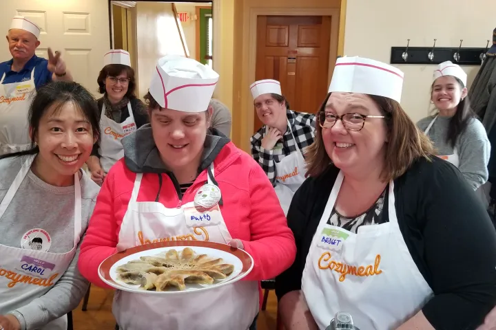 Group of people in aprons and hats smiling with a plate of food during a cooking class.