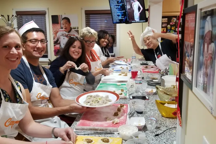 Six people cooking and smiling at a kitchen counter, wearing aprons and chef hats.