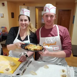 Two people in aprons holding a plate of dumplings in a kitchen setting.