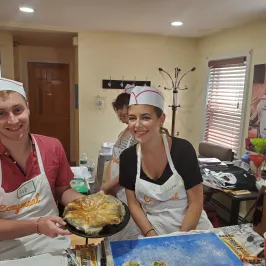 Two smiling people in chef hats and aprons holding a baked dish in a kitchen setting.
