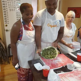 Three people wearing aprons hold a plate of green pasta in a kitchen setting.