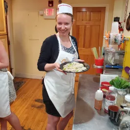 Person in apron and hat holding a plate of food in a kitchen.