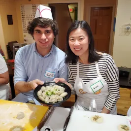 Two people smiling, holding a plate of dumplings in a kitchen setting.