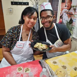 Two people smiling, holding a plate of dumplings, with more being prepared on the table.