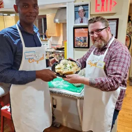Two people in aprons hold a plate of food in a kitchen with an exit sign in the background.