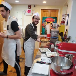 People in a kitchen wearing aprons and hats, with one holding a tray of food and cups.