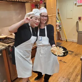 Two women in aprons and chef hats posing with a plate of food in a kitchen.