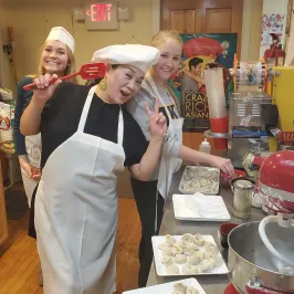 Three people in a kitchen wearing aprons, preparing dumplings with a mixer on the counter.
