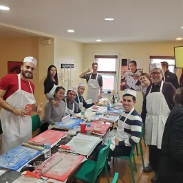 Group of people wearing aprons and hats, gathered around tables for a cooking class in a cozy room.