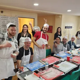Group of people in aprons and hats enjoying a cooking class in a room with tables and ingredients.