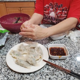 Person making dumplings on a kitchen counter with a plate of dumplings and dipping sauce.