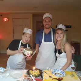 Three people in aprons and chef hats in a kitchen, posing with food on a table.