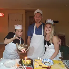 Three smiling people in aprons and chef hats with a decorative pot near a table of prepared food.