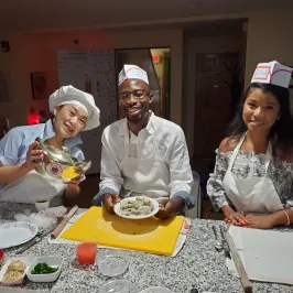 Three people wearing aprons smiling at a kitchen counter with food.