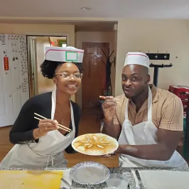 Two people in aprons and paper hats holding a caramel-patterned dessert plate with chopsticks.