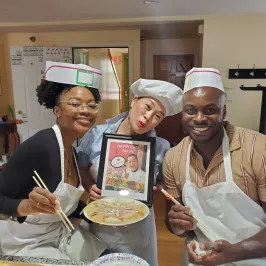 Three people smiling with aprons and hats, holding a framed photo and a plate with chopsticks.