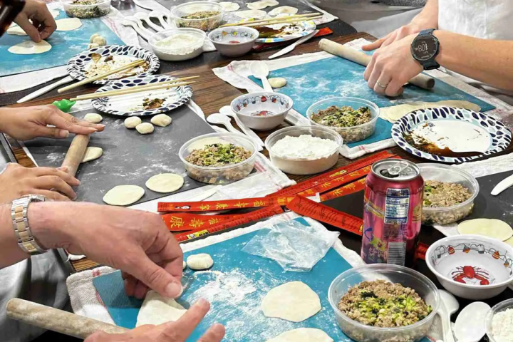 Group of people making dumplings with fillings at a table, using rolling pins and bowls of ingredients.