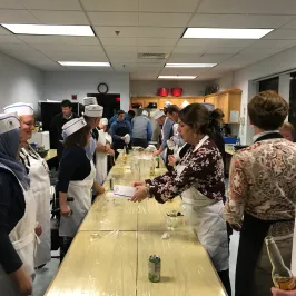 People in chef hats and aprons gathered around tables in a kitchen setting.