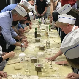 People in chef hats making food at a long table, rolling dough with rolling pins.
