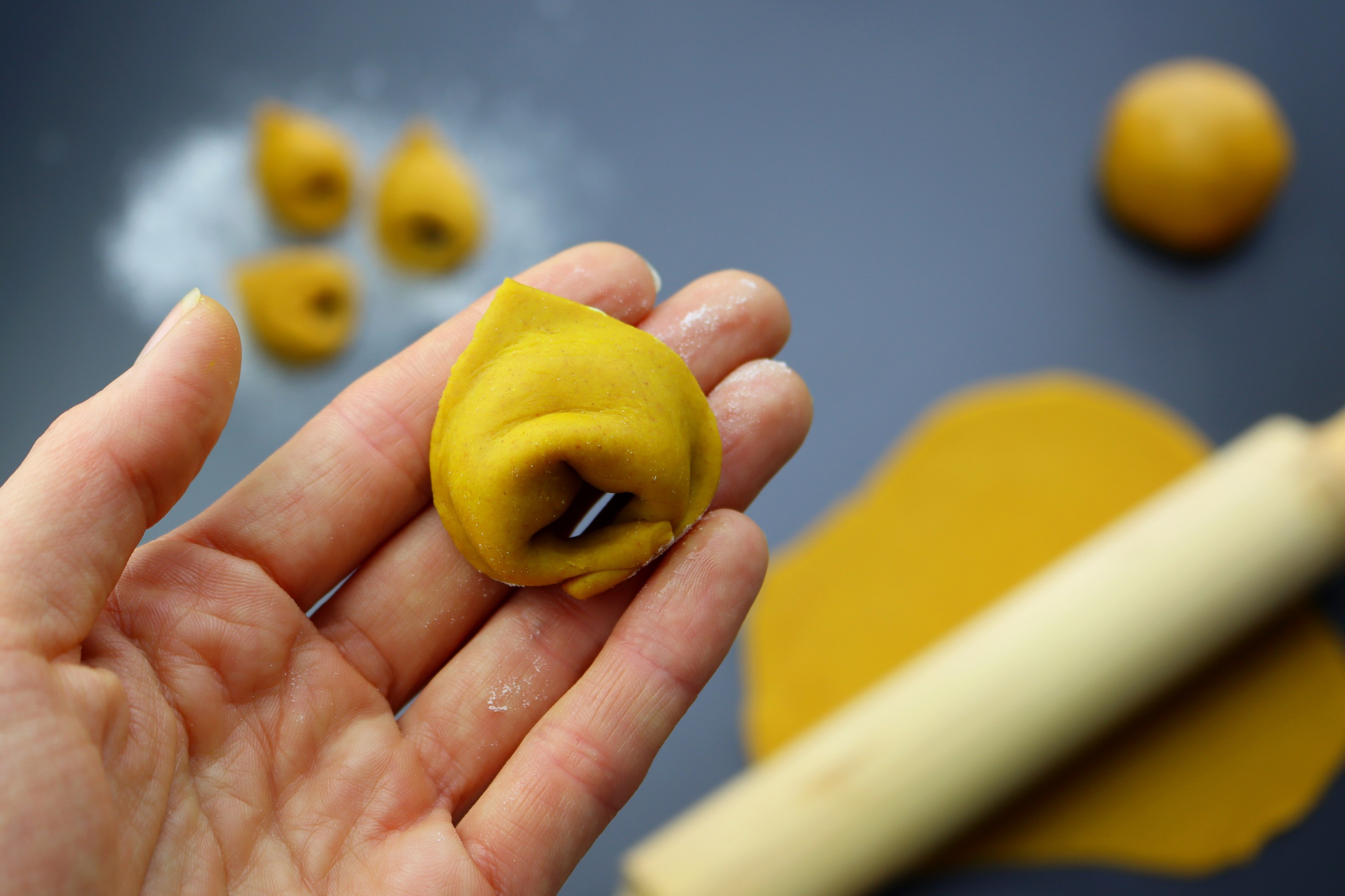 Hand holding tortellini with more dough and a rolling pin on a table.